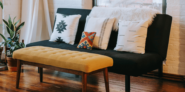 Sofá con cojines étnicos y banco mostaza en salón cálido, representación del interiorismo atemporal en muebles que invitan al descanso y la conexión.