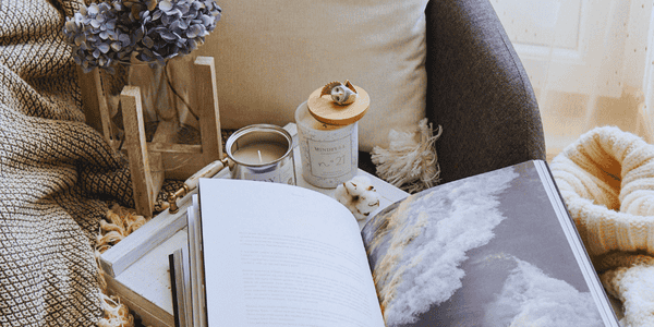 Rincón de lectura acogedor con libro abierto, manta suave, velas aromáticas y decoración en tonos cálidos sobre un sillón gris, ideal para crear un espacio de calma y autocuidado en casa.